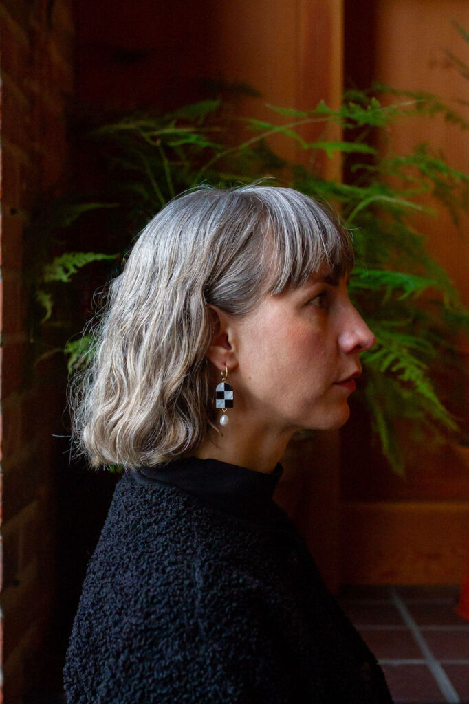 Side profile of woman with grey hair wearing black and white checkered earrings and plants in background